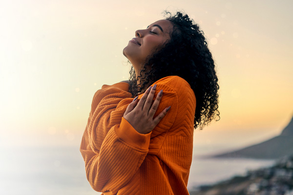 A woman in an orange sweater hugging herself at sunset